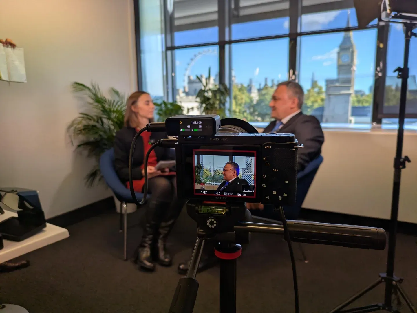 A filming camera frames a host-led interview with Westminster visible beyond the window.
